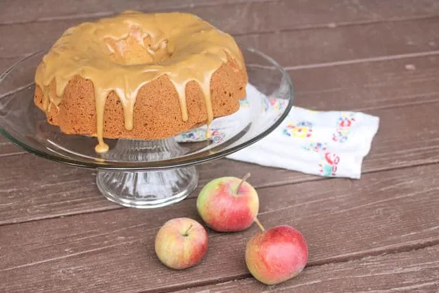 A bundt cake with frosting dripping down the sides sitting on a glass cake plate surrounded by fresh apples.