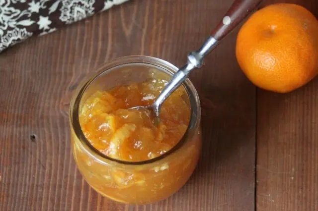 An open jar of clementine rum marmalade with a spoon inside as seen from above, a fresh clementine and napkin in the background.