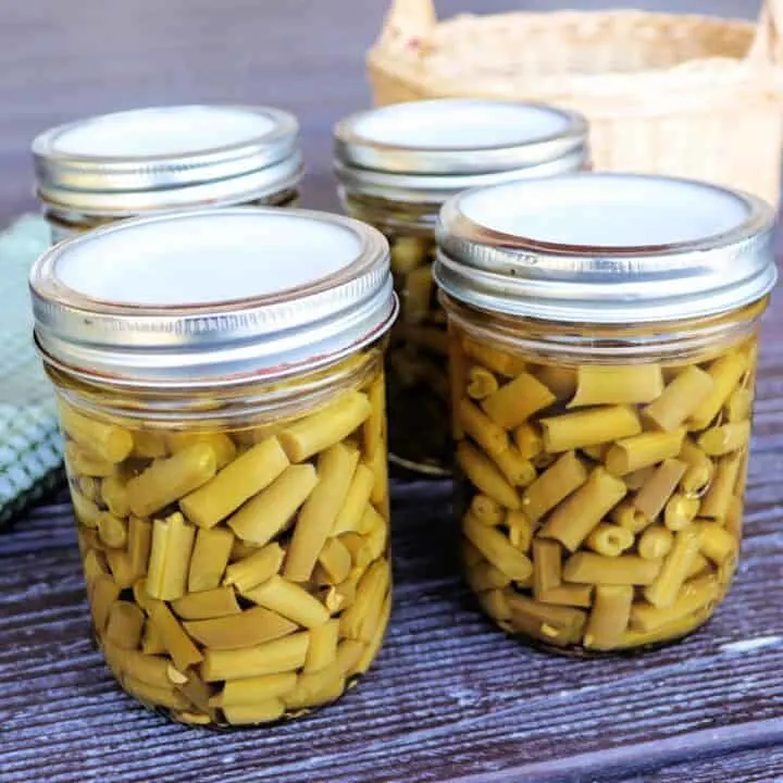 Jars of green beans sit on a table with a basket in the background.