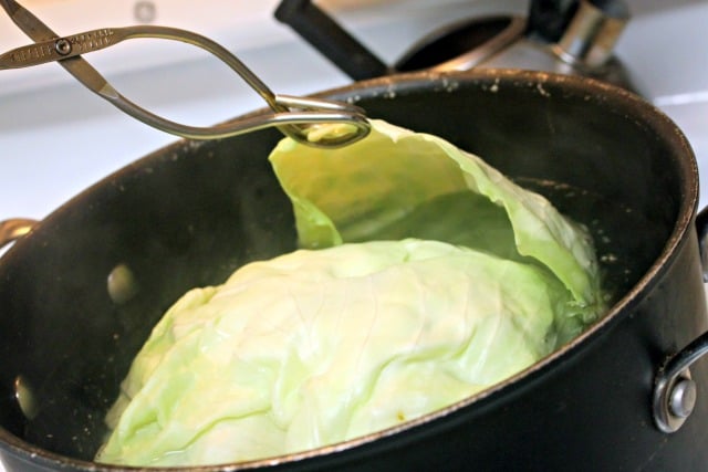 A pair of metal tong separating a cabbage leaf from the entire head in a pot of boiling water.