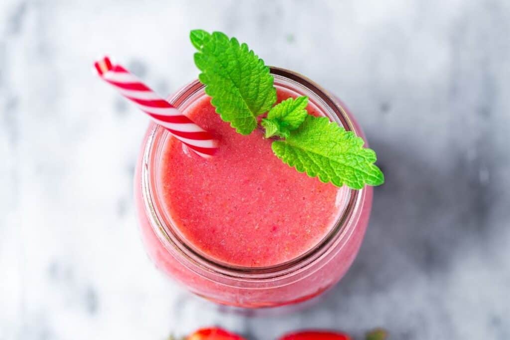 A glass full of red smoothie with a straw and fresh lemon balm leaves as seen from above.