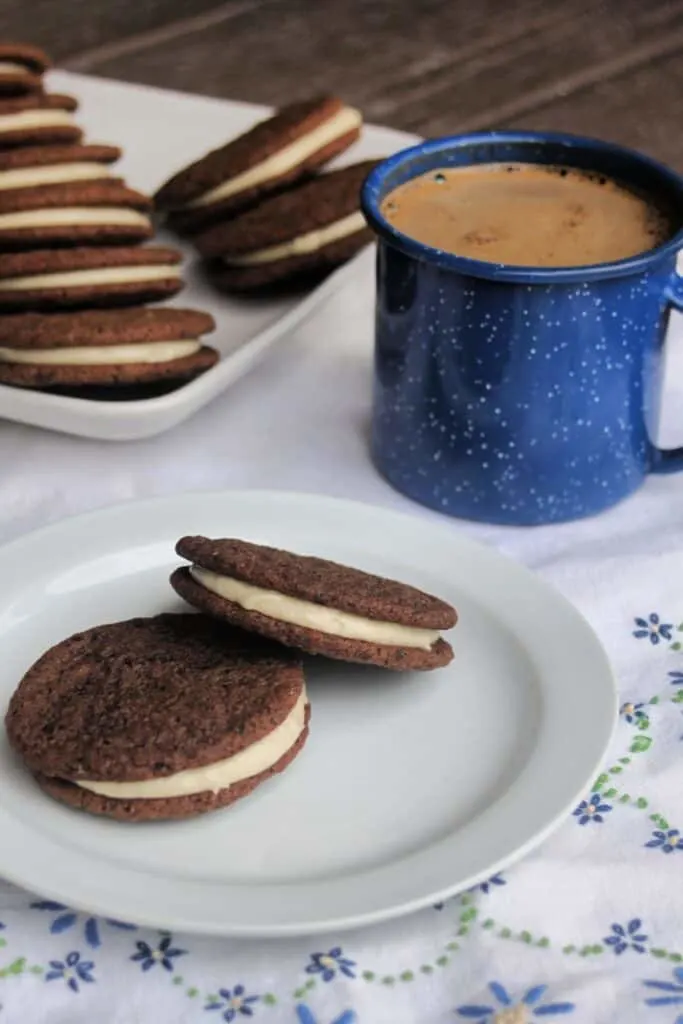 2 chocolate sandwich cookies sitting on a plate, with a platter of more cookies in the background and a blue tin cup full of coffee.