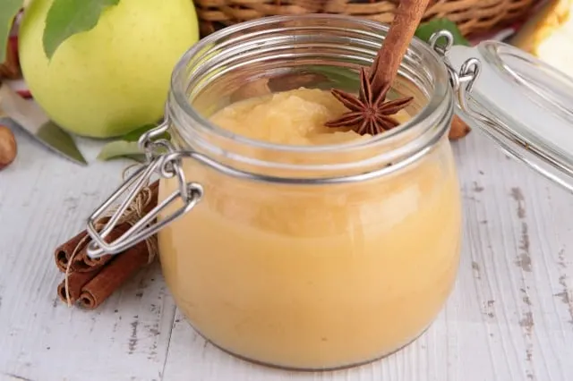 Jar of applesauce surrounded by fresh apples and cinnamon sticks.