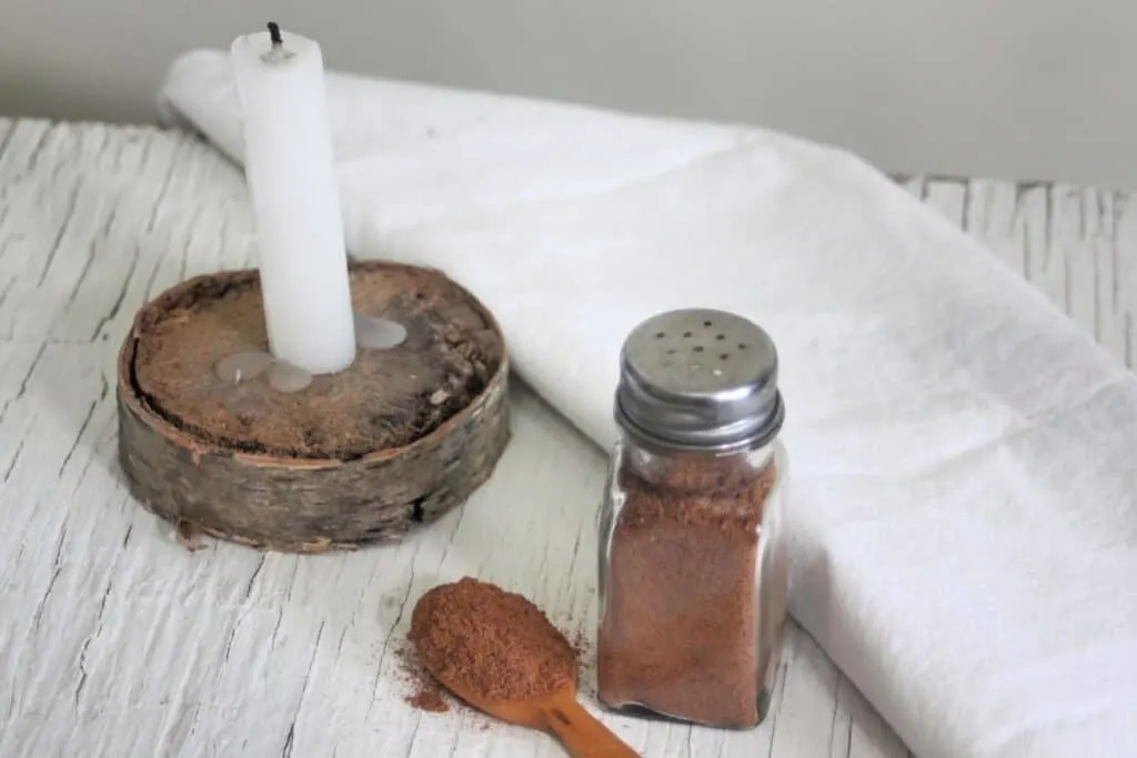 A wooden spoon full of ground flaxseed and spices sitting next to a salt shaker full of the same mixture with a candle and white linen napkin on a table.
