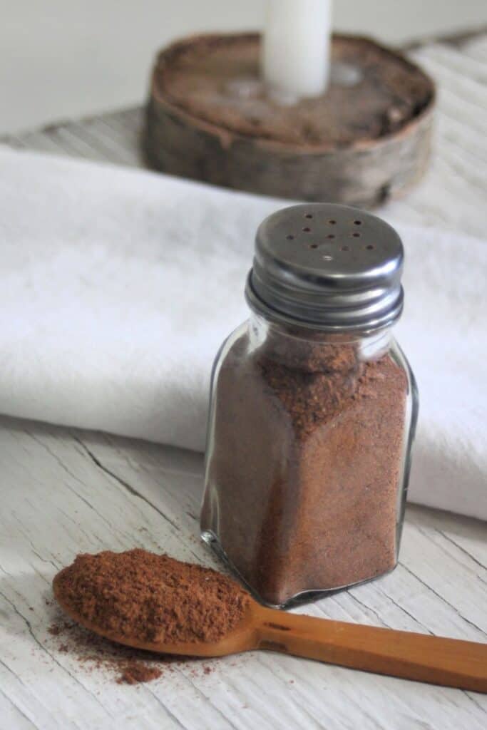 A wooden spoon full of ground flaxseed and spices sitting next to a salt shaker full of the same mixture with a candle and white linen napkin on a table.