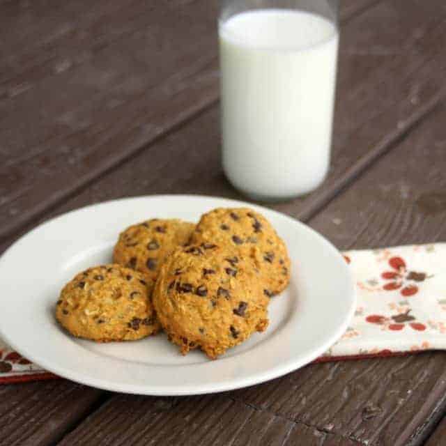Pumpkin chocolate chip oatmeal cookies on a plate that is sitting on top of a napkin with a glass of milk in the background.