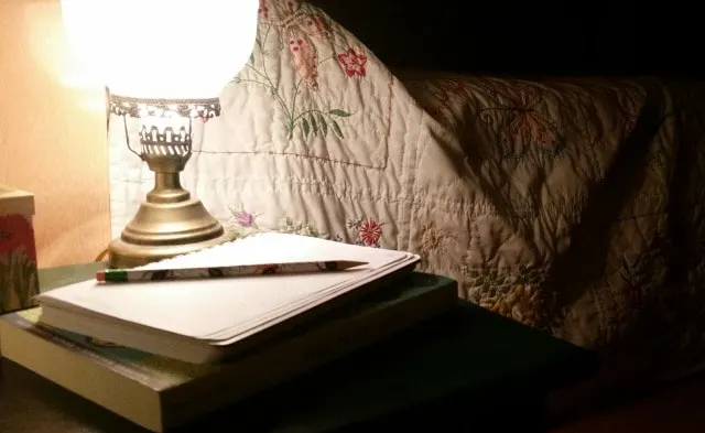 A lamp lit up beside a quilt covered bed in a dark room.