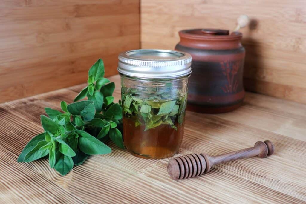 A sealed jar of honey with green leaves floating in it sits on a table. Next to it is a stem of fresh oregano leaves and a wooden honey dipper. In the background is a clay honey pot.