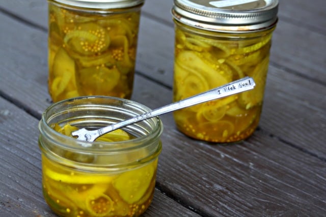 An open jar of bread and butter pickles with a pickle fork stuck inside sitting in front of 2 sealed jars of pickles.