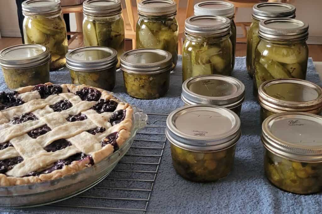 Jars of pickles are sitting on a towel on a kitchen counter. A pie is also cooling on a wire rack next to the jars. 