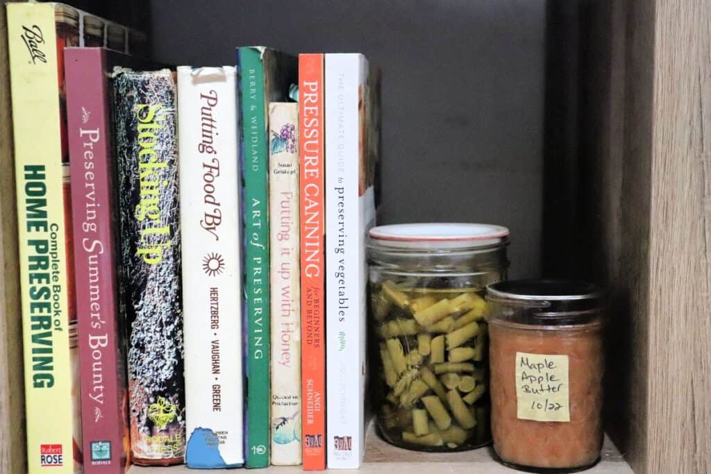 Canning books lined up on a bookshelf with a jar of canned green beans and apple butter sitting next to the books.