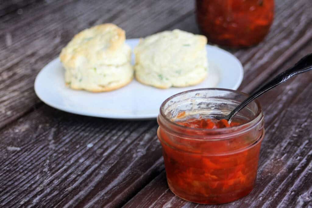 An open jar of an orange colored jam sits on a table with a plate of biscuits sitting behind it. 