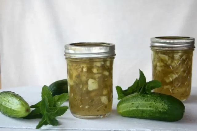 Jars of cucumber mint jam on a table with whole cucumbers and fresh mint.