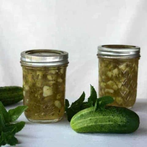 Jars of cucumber mint jam on a table with whole cucumbers and fresh mint.