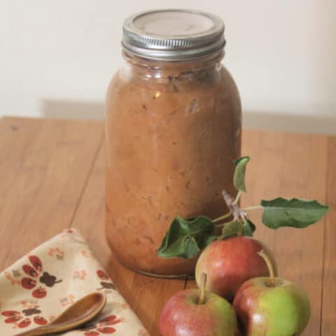 A jar of chunky applesauce on a table with fresh apples, a napkin, and a spoon.