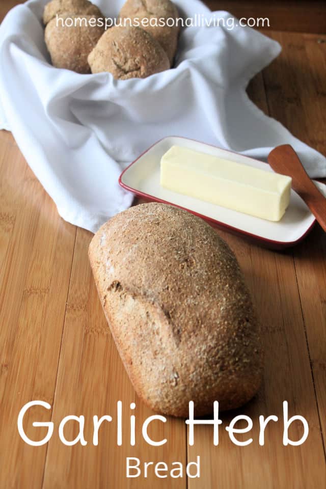 A loaf of garlic and herb bread on a board with a stick of butter and rolls in a napkin lined basket.