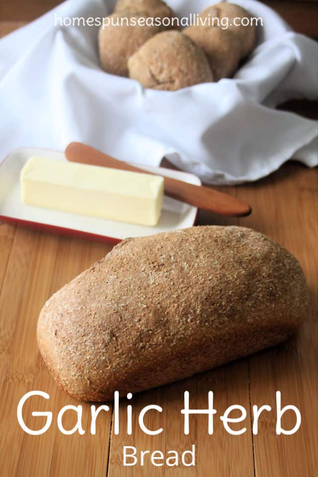 A loaf of garlic and herb bread on a board with a stick of butter and rolls in a napkin lined basket.