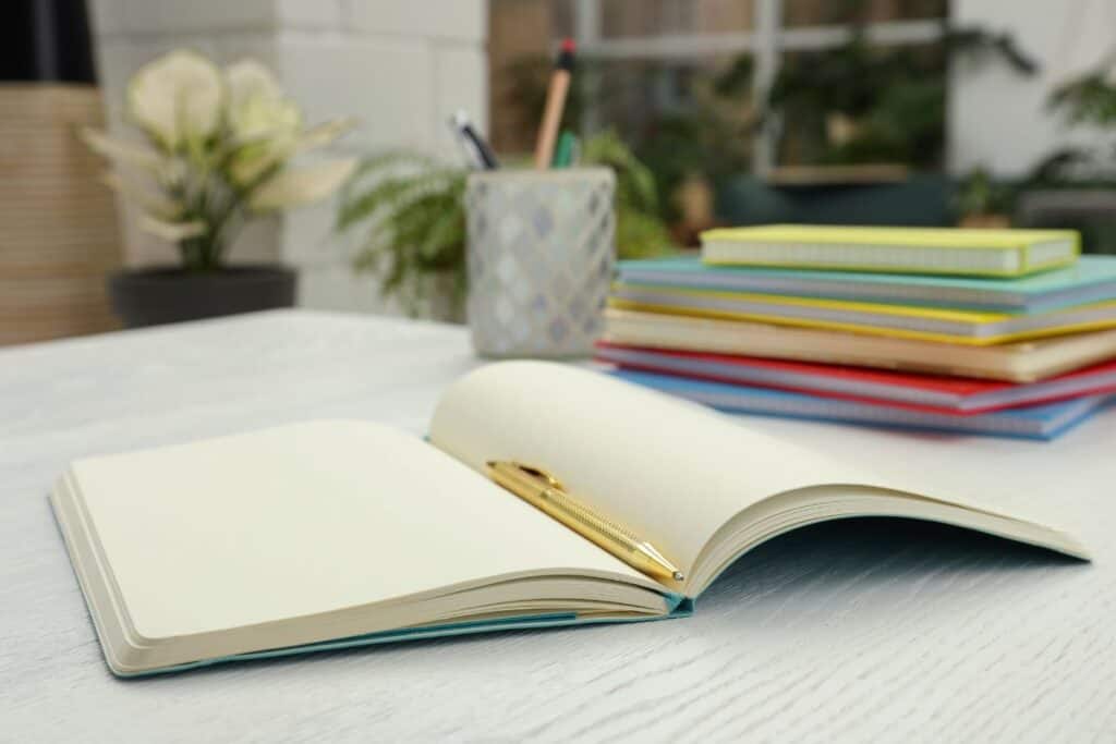 An open book with blank pages sits with a pen on top on a table. A stack of journals a cup with pens inside sits in the background.