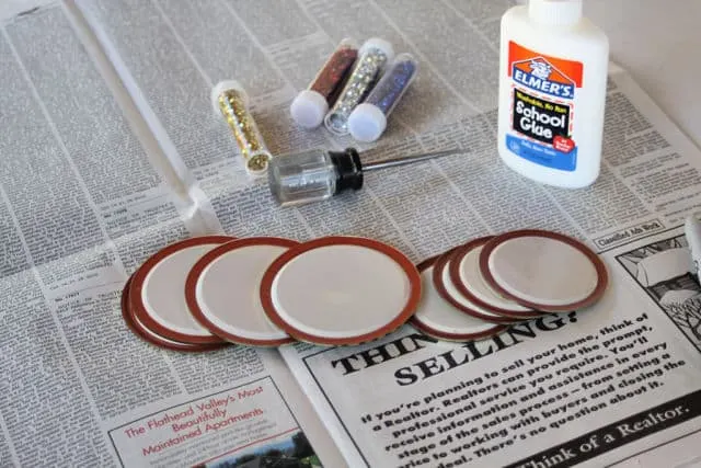 Canning lids, glue, an awl, and glitter spread out on newspaper to make upcycled canning lid gift tags.
