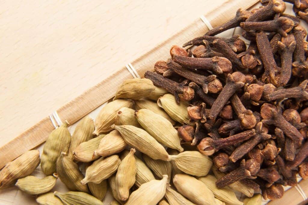 Whole cardamom pods sit next to whole cloves on a board. 