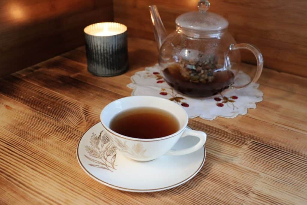 A tea cup and saucer sit in front of a clear glass tea pot and candle.