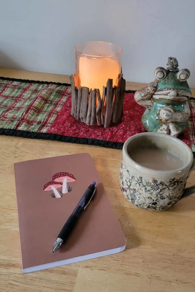 A closed journal with a pen on top sits on a table with a cup of coffee and lit candle on a table runner in the background.