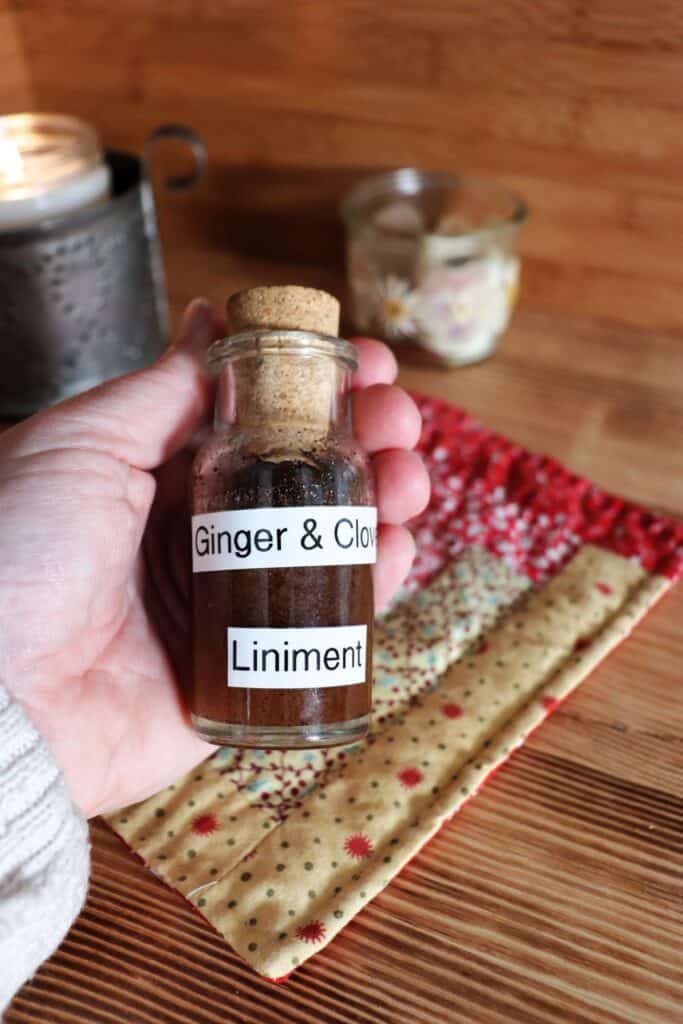 A woman's hand holds a small glass bottle with a dark liquid, labeled ginger and clove liniment. Candles and cloth sit on the table in the background. 