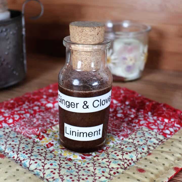 A small glass bottle with a cork lid full of dark liquid sits on a quilted square with candles in the background. The glass bottle is labeled Ginger & Clove Liniment.