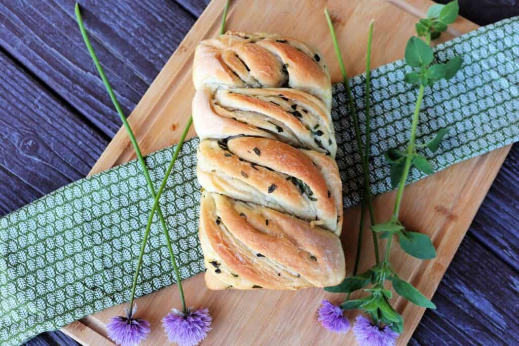 A swirled loaf of bread with herbs in the crust is seen from above. It sits on a board surrounded by fresh herbs.