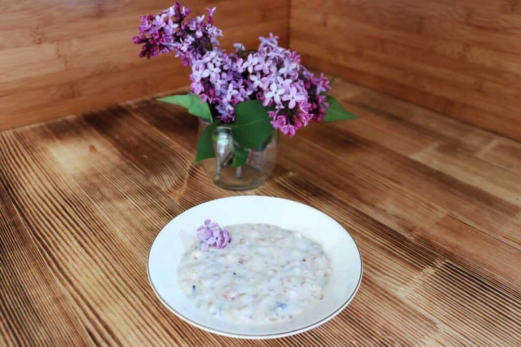 A bowl of rice pudding sits on a table garnished with fresh lilac flowers. A vase of fresh lilac blooms sits behind it.