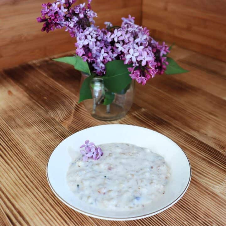 A bowl of rice pudding that is garnished with lilac flowers sits on a table. A vase full of lilac branches sits in the background.
