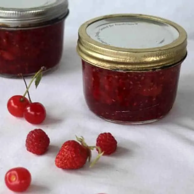 Jars of brandied cherry berry preserves on a table with fresh berries and cherries.