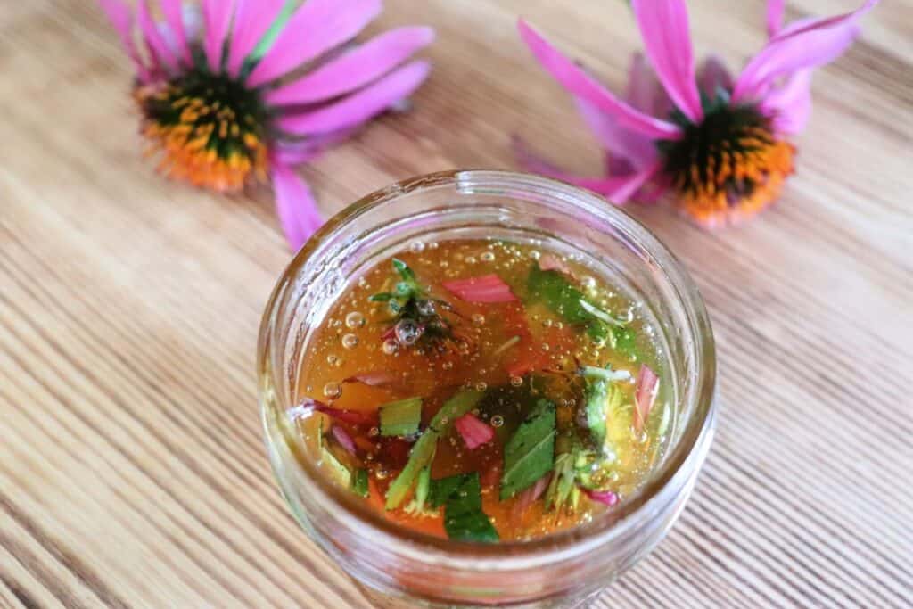 An open glass jar full of honey and purple flowers and green leaves sits on a table. Behind it sits two purple coneflowers in bloom. 