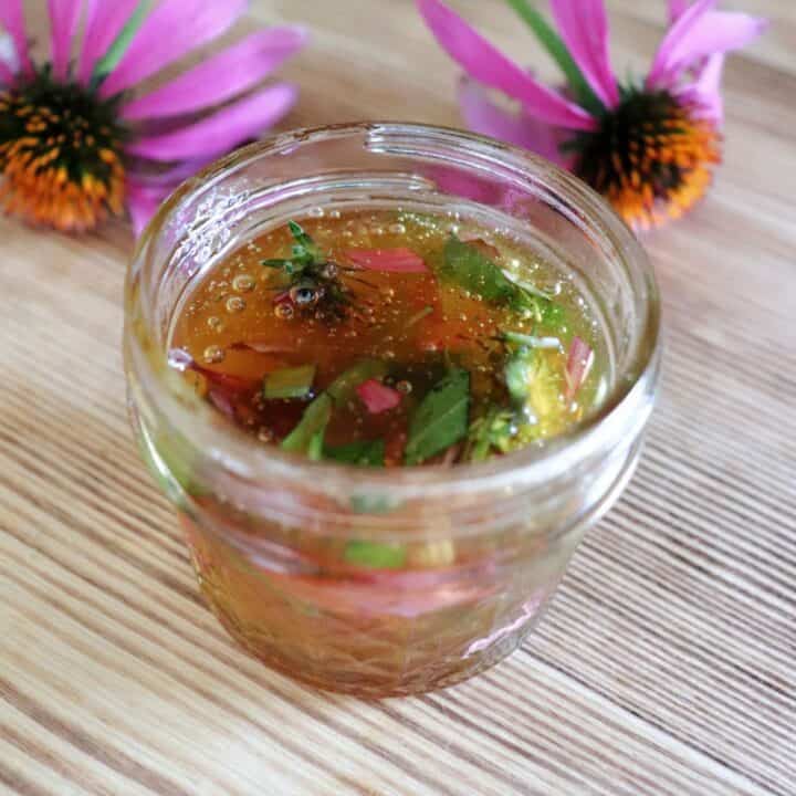 An open small glass jar full of honey and chopped purple flowers and green leaves sits on a table. Behind the jar sits two blooming purple coneflowers on their stems.