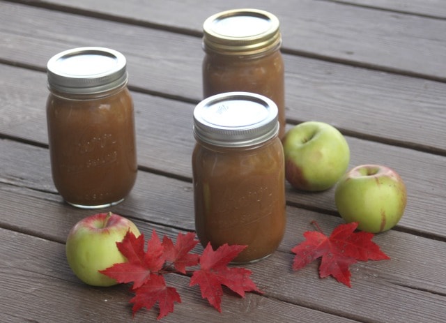 Jars of maple apple butter on a table surrounded by red maple leaves and fresh apples. 