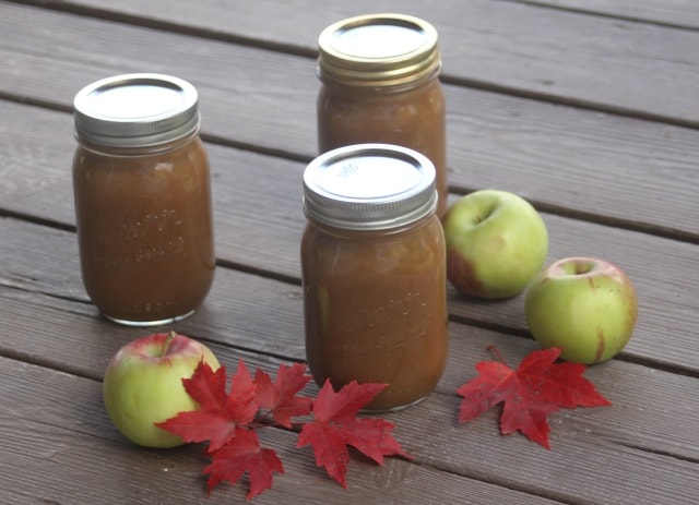 3 jars of maple apple butter on wooden boards with fresh apples and red maple leaves.