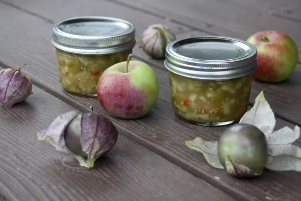 Jars of apple salsa verde on a table surrounded by fresh apples and tomatillos.