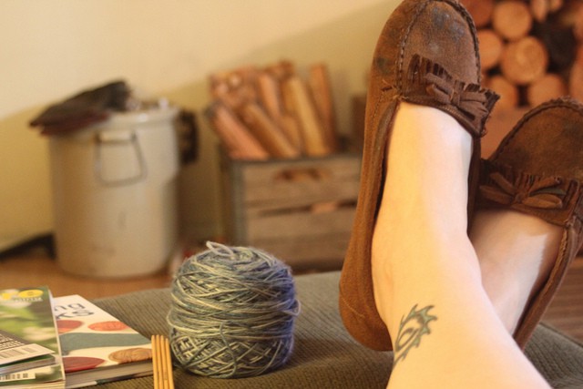 Legs and feet in slippers on a footstool with a ball of yarn and knitting needles. 