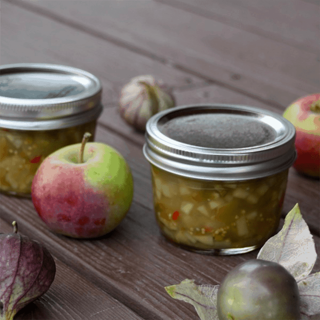 Jars of Apple Salsa Verde on a table surrounded by fresh apples and tomatillos.