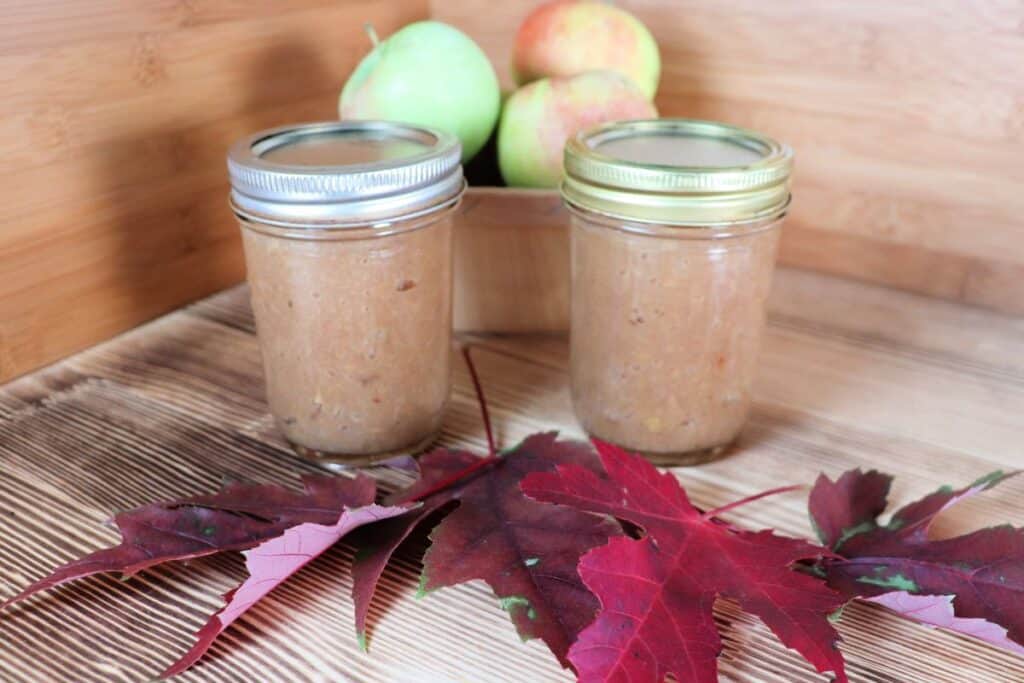 Two sealed jars of apple butter sit in front of a basket of apples. In front of the jars are red maple leaves.