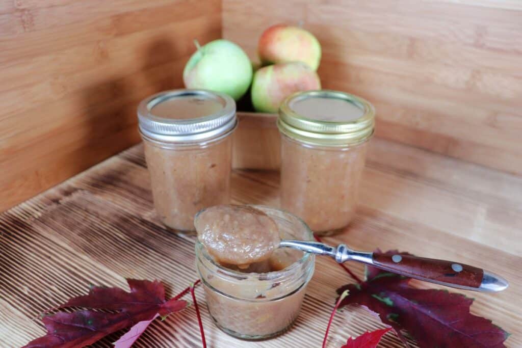 An open jar of apple butter with a spoon sitting on top sits on a table. Behind it are more sealed jars of apple butter and a basket of fresh apples. In front are red maple leaves. 
