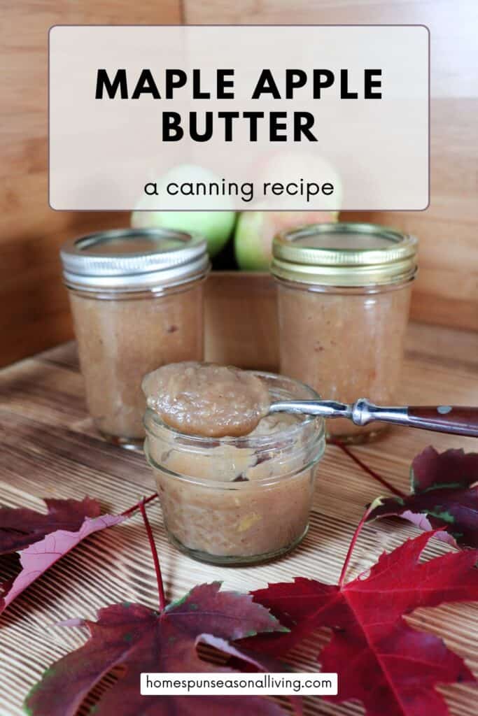An open jar of apple butter with a spoon sitting on top sits on a table. Behind it are more sealed jars of apple butter and a basket of fresh apples. In front are red maple leaves. Text overlay reads: Maple Apple Butter - a canning recipe. 
