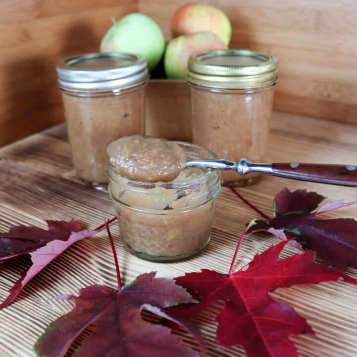 An open jar of apple butter with a spoon sitting on top sits on a table. Behind it are more sealed jars of apple butter and a basket of fresh apples. In front are red maple leaves.
