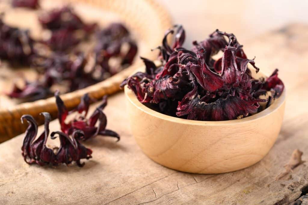 A small wooden bowl full of dried hibiscus roselles sits on a table with more roselles next to and behind the bowl on the table. 