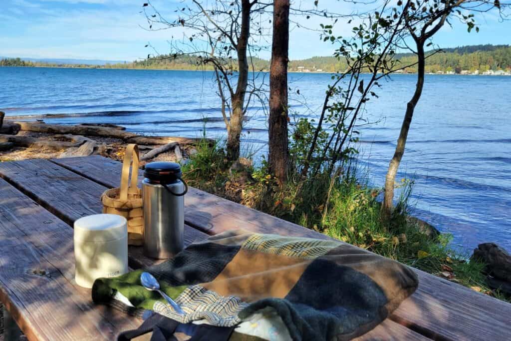 A patchwork bag, metal containers, and a basket sit on a picnic table in front of a lake. 