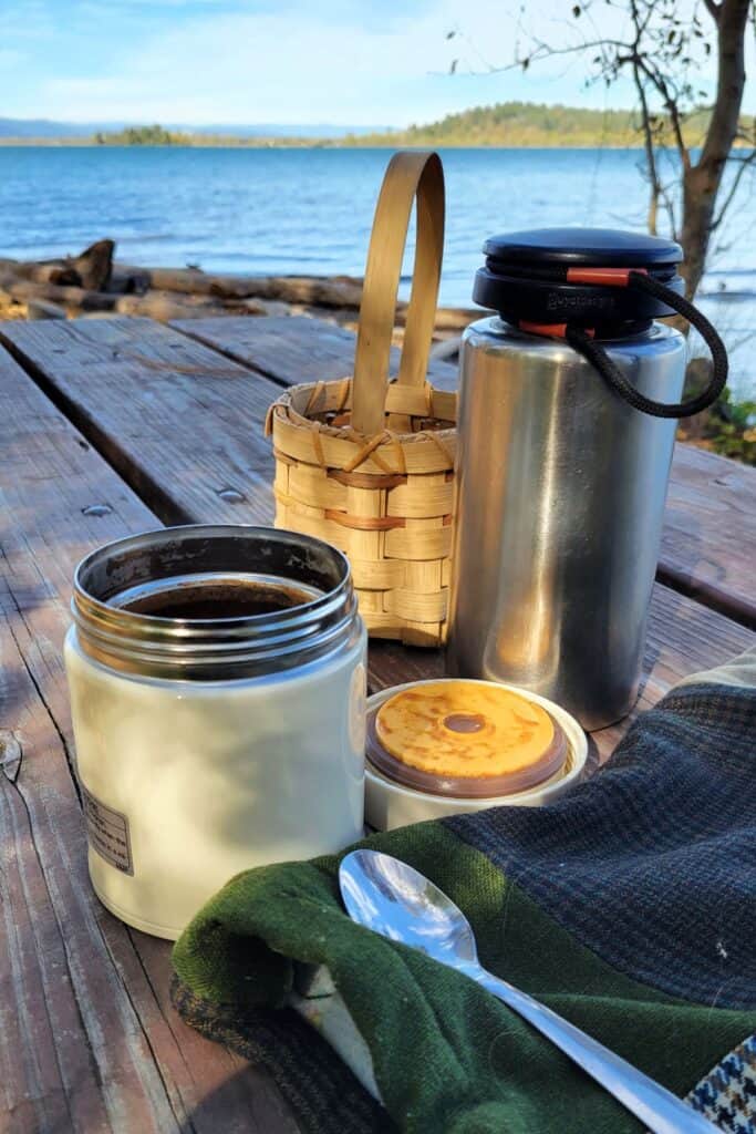 Metal containers sit on a picnic table in front of a lake. A spoon sits on a cloth next to the containers.