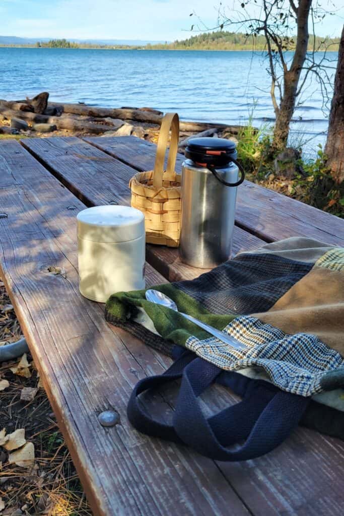 A patchwork bag, metal containers, and a basket sit on a picnic table in front of a lake. 