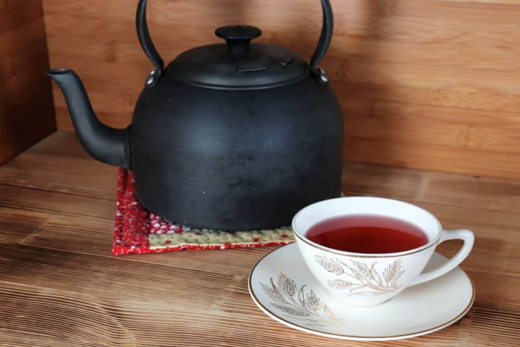 A teacup sits on a saucer with a metal teapot on a red cloth sits in the background. The tea cup is full of a red herbal tea.