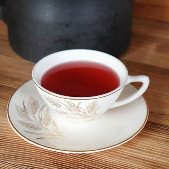 A teacup sits on a saucer. The teacup is full of a red tea. A metal teapot sits in the background.