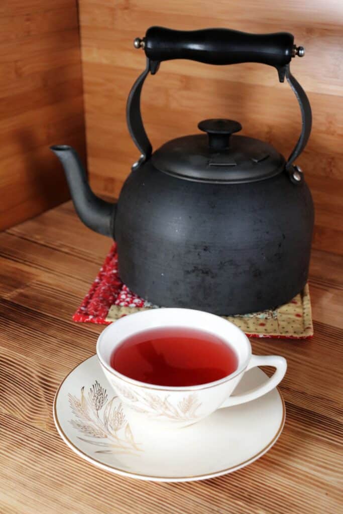 A teacup sits on a saucer with a metal teapot on a red cloth sits in the background. The tea cup is full of a red herbal tea.